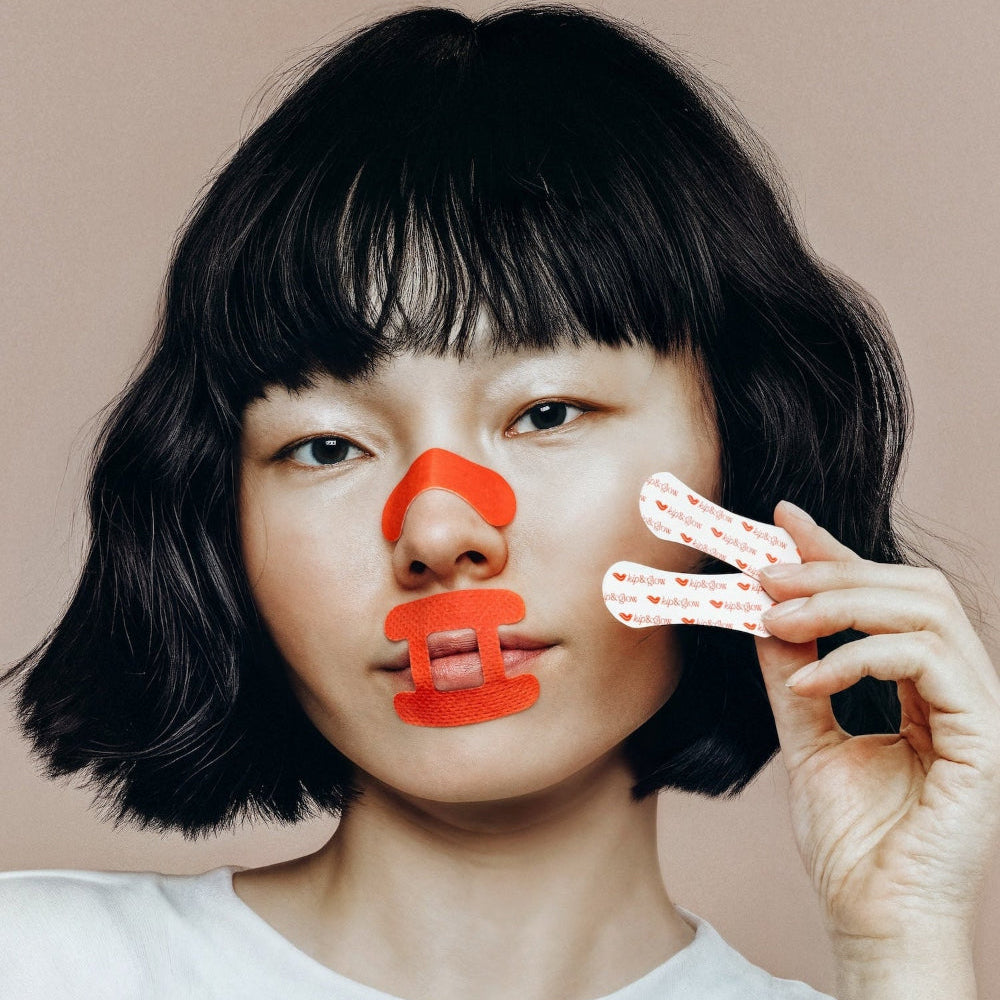 Woman with bobbed dark hair holding up kip&glow nose strip whilst wearing the sleep tape and nose strip