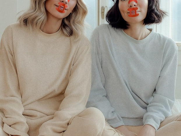 Two people sitting on a bed in a bright room with large windows, wearing kip&glow nose strips and sleep tape.